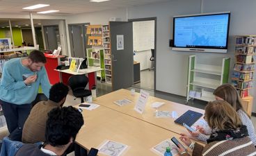En gruppe som har IKT-kurs i biblioteket. De sitter rundt et bord og ser på mobiler og en storskjerm.