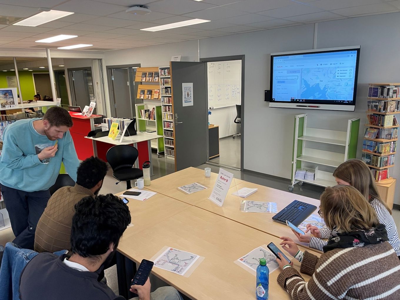 En gruppe som har IKT-kurs i biblioteket. De sitter rundt et bord og ser på mobiler og en storskjerm. En gruppe som har IKT-kurs i biblioteket. De sitter rundt et bord og ser på mobiler og en storskjerm.